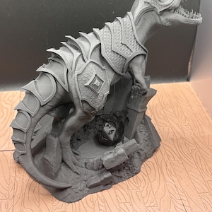 Puede incluir: Una figura gris impresa en 3D de un Tyrannosaurus Rex con armadura, de pie sobre una base rocosa. Un dado de múltiples caras descansa en una pequeña cueva. El dinosaurio tiene escamas y armadura detalladas. La base tiene un patrón de vetas de madera.