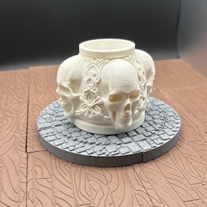 Puede incluir: Un recipiente crema con cuatro calaveras en los lados. Se apoya sobre una base gris circular con un diseño de adoquines. El fondo es marrón oscuro.