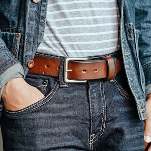 May include: A brown leather belt with a silver buckle. The belt is worn over a pair of blue jeans and a denim jacket.