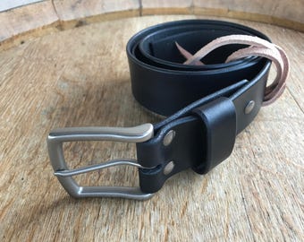 Op de afbeelding: Een zwarte leren riem met een zilveren gesp. De riem is gevouwen en de gesp is zichtbaar. De riem ligt op een houten oppervlak.