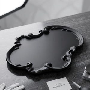 May include: A black decorative tray with an ornate, baroque-style design. The tray has a smooth, glossy surface and a detailed, scrolled edge. A small card with the text "MODEL: BAROQUE-F1" is visible.