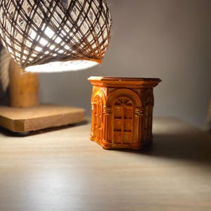 May include: An orange, rectangular decorative planter with arched window designs. The planter is illuminated by a hanging lamp with a woven shade. The lamp is suspended above a wooden surface, creating a warm, inviting ambiance.