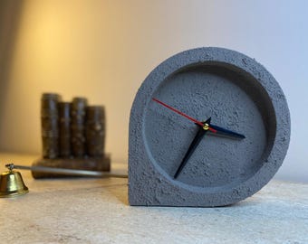 May include: A gray concrete clock with black hands and a red second hand. The clock has a rounded top and a textured surface. A small brass bell and a wooden barrel decoration are also visible.