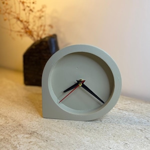 May include: A light gray, minimalist desk clock with black hands and a red second hand. The clock has a unique, rounded design and sits on a textured surface. A dark vase with dried flowers is in the background.