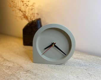May include: A light gray, minimalist desk clock with black hands and a red second hand. The clock has a unique, rounded design and sits on a textured surface. A dark vase with dried flowers is in the background.