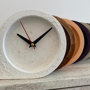 May include: A contemporary white concrete clock with black hands and a red second hand. The clock face is round, and the clock has a textured surface. A row of colourful, circular objects in shades of brown, orange, purple, and grey are behind the clock.
