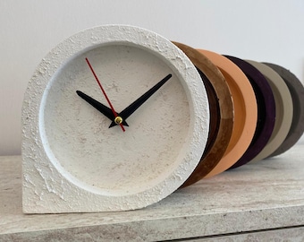 May include: A modern, white concrete clock with black hands and a red second hand. The clock face is round, and the clock has a textured surface. A row of colorful, circular objects in shades of brown, orange, purple, and gray are behind the clock.