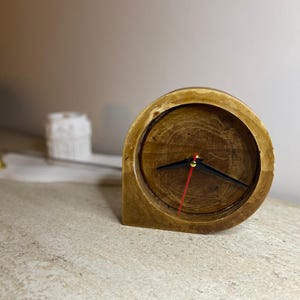 Puede incluir: Un pequeño reloj redondo de madera con un acabado marrón claro. El reloj tiene manecillas negras y un segundero rojo. El reloj está sobre una superficie de color claro.