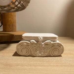 Pode incluir: Pequena caixa decorativa esbranquiçada com um design clássico ornamentado. A caixa apresenta um topo retangular e uma base detalhada com motivos florais e de volutas. A caixa está sobre uma superfície de madeira clara.