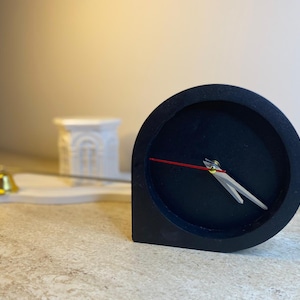 May include: A black, minimalist desk clock with a round face and a red second hand. The clock has white hands and a unique, geometric shape. A small gold bell and a white decorative piece are in the background.