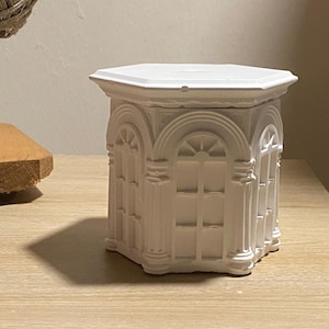 May include: A white, hexagonal-shaped decorative box with architectural details. The box features arched windows and columns, creating a classical design. The box is made of a solid material and sits on a light-colored surface.