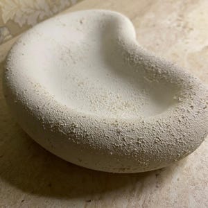 May include: A white, kidney-shaped ceramic bowl with a textured, speckled surface. The bowl has a rounded rim and a shallow interior. The colour is a muted off-white, with a slightly rough texture.