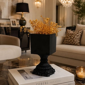 Pode incluir: Um vaso quadrado preto com base de pedestal contém um ramo de flores amarelas secas. O vaso repousa sobre dois livros empilhados, o superior intitulado CEREAL e o inferior intitulado LIVE BEAUTIFULLY de Calderone, sobre uma superfície escura.