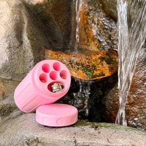 May include: A pink cylindrical container with a removable lid, designed to hold multiple small items. The container is open, revealing several compartments and a metal object. The lid is circular with two recessed circles. The background features a natural rock formation and flowing water.