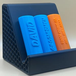 May include: A blue and orange charging stand with two slots for charging devices. The stand is made of a textured black plastic. The slots are labeled "DAVID" and "GROOMSMEN".