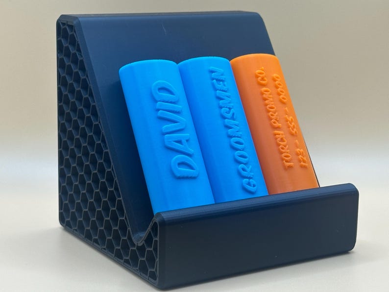 May include: A blue and orange charging stand with two slots for charging devices. The stand is made of a textured black plastic. The slots are labeled "DAVID" and "GROOMSMEN".