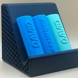 May include: A dark blue, hexagonal patterned stand holds three blue and turquoise cylindrical containers. The containers are labeled "DAVID" and "GROOMSMEN".