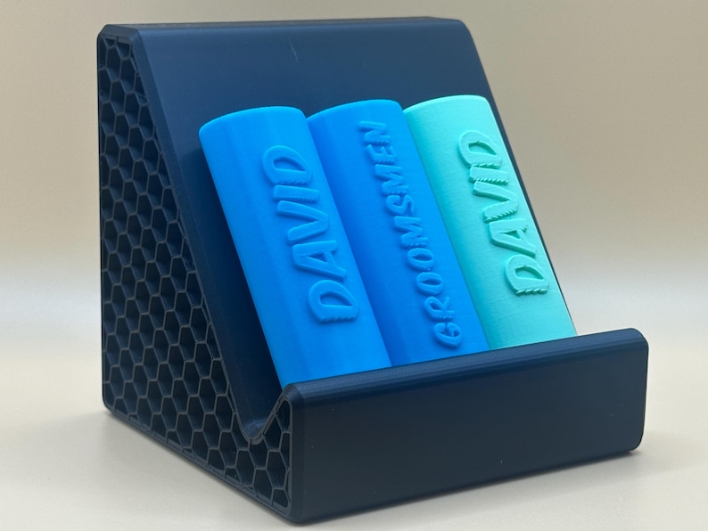 May include: A dark blue, hexagonal patterned stand holds three blue and turquoise cylindrical containers. The containers are labeled "DAVID" and "GROOMSMEN".