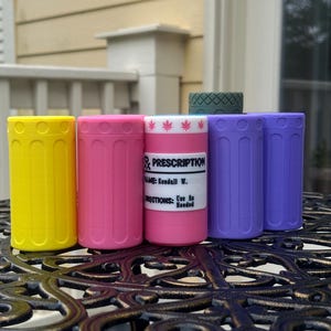 Könnte beinhalten: Eine Sammlung farbenfroher, zylindrischer Behälter in Gelb, Pink, Lila und eine Medikamentenflasche mit Etikett. Die Behälter sind auf einem dekorativen Metalltisch angeordnet, mit einem grünen Behälter im Hintergrund. Die Medikamentenflasche hat ein Etikett mit Text.