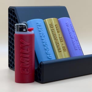 Op de afbeelding: Een zwarte plastic aanstekerhouder met drie vakken. De houder heeft een honingraatpatroon aan de zijkanten. Er zitten drie aanstekers in de houder: een rode aansteker met de naam "EMILY" erop gedrukt, een blauwe aansteker met de tekst "GROOMSMEN" erop gedrukt en een gouden aansteker met de tekst "122 - 355 - 00000  TOCCI PRINT CO" erop gedrukt.