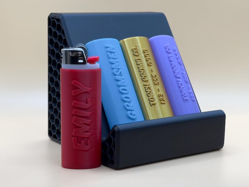May include: A black plastic lighter holder with three slots. The holder has a honeycomb pattern on the sides. There are three lighters in the holder: one red lighter with the name "EMILY" printed on it, one blue lighter with the text "GROOMSMEN" printed on it, and one gold lighter with the text "122 - 355 - 00000  TOCCI PRINT CO" printed on it.