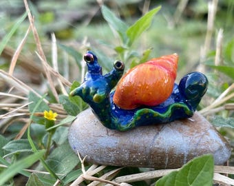 May include: A handmade ceramic snail ornament. The snail has a blue body and an orange shell, with large eyes. It sits on a small, grey stone, amidst green grass and tiny yellow flowers. A whimsical garden decoration.