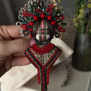 Può includere: Un broche di perline con un designo rosso, verde e oro. Il broche presenta una figura umana stilizzata con una corona di perline e un lungo ciondolo di perline.