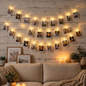 May include: A decorative string of fairy lights with small clips holding photographs, strung across a white wooden wall. Below the lights is a beige sofa, framed pictures, and candles, creating a cosy atmosphere.