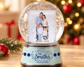 Puede incluir: Una bola de nieve de cristal transparente que contiene una fotografía rectangular de una familia de cuatro personas. La base es azul claro con puntos azul oscuro y el texto THE SMITHS en una fuente cursiva decorativa.