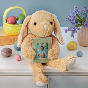 May include: A plush tan bunny holding a green framed photo of a child wearing bunny ears. Easter eggs in a yellow basket and scattered around the bunny. A vase of purple flowers sits to the right.