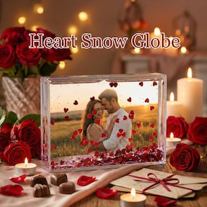 May include: A rectangular snow globe with a photo of a couple embracing, surrounded by red heart-shaped confetti. The words "Heart Snow Globe" are displayed above the globe. Red roses and lit candles are in the background.
