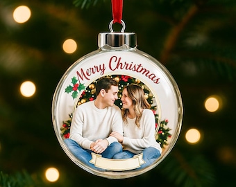 Op de afbeelding: Transparante glazen kerstbal met een rood lint. De ornament bevat een foto van een stel en de woorden "Merry Christmas" in het rood. Een takje hulst staat aan de linkerkant van de ornament.