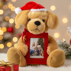 Puede incluir: Un perro de peluche marrón dorado con un gorro de Papá Noel rojo, sosteniendo un marco de fotos rojo. El marco muestra un retrato familiar frente a un árbol de Navidad. El perro tiene una nariz negra y está sentado sobre una superficie blanca.