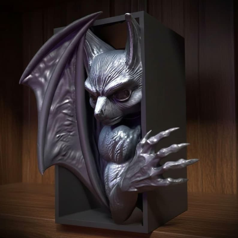 May include: A dark gray, gothic-style sculpture of a bat-like creature emerging from a black rectangular box. The sculpture features detailed wings, a stern face, and clawed hands, creating a dramatic and imposing aesthetic. The sculpture is likely a decorative item.