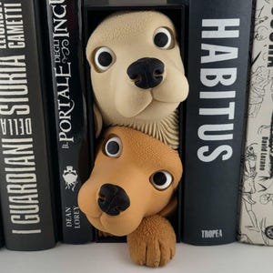 May include: A whimsical dog-shaped bookend set in cream and brown, peeking out from between black book spines. The bookend features large, expressive eyes and a textured appearance, adding a playful touch to any bookshelf. The word "HABITUS" is visible on one of the book spines.
