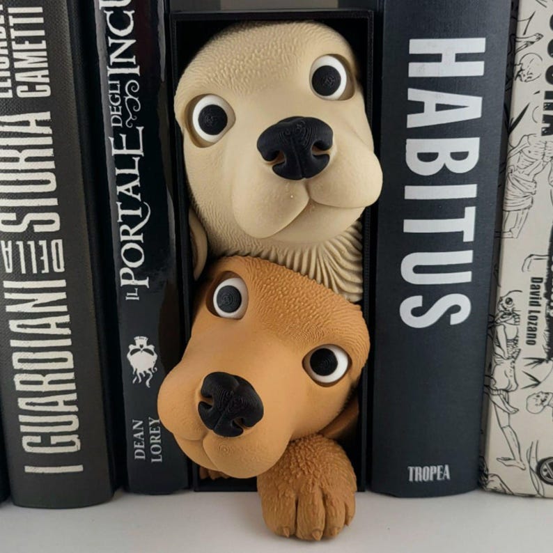 May include: A whimsical dog-shaped bookend set in cream and brown, peeking out from between black book spines. The bookend features large, expressive eyes and a textured appearance, adding a playful touch to any bookshelf. The word "HABITUS" is visible on one of the book spines.