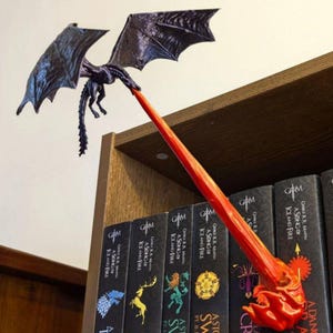 May include: A black dragon-shaped bookmark with outstretched wings, appearing to breathe a red flame. The bookmark is placed among a row of books with the text "A Song of Ice and Fire" on the spines. The dragon is perched on a wooden shelf.