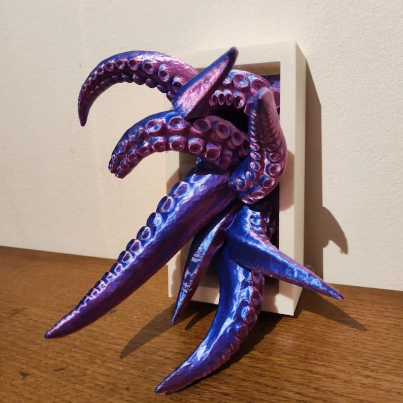 May include: A 3D-printed sculpture of purple and blue tentacles emerging from a white rectangular frame. The tentacles have a textured surface and are arranged to appear as if they are reaching out. The sculpture is placed on a wooden surface.