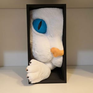 May include: A white cat face sculpture with a bright blue eye and orange nose, framed in a black rectangular box. The cat's paw extends out of the frame, adding a playful touch to the artwork.