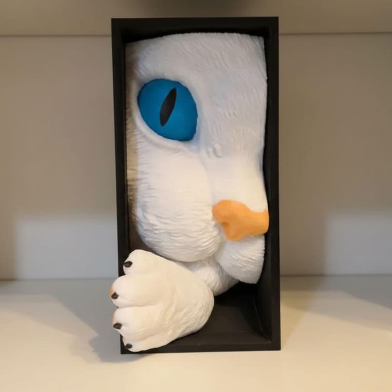 May include: A white cat face sculpture with a bright blue eye and orange nose, framed in a black rectangular box. The cat's paw extends out of the frame, adding a playful touch to the artwork.