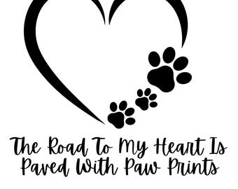 May include: Black graphic of a heart with paw prints trailing from it. The text "The Road To My Heart Is Paved With Paw Prints" is below the heart and paw prints. The image is on a white background.