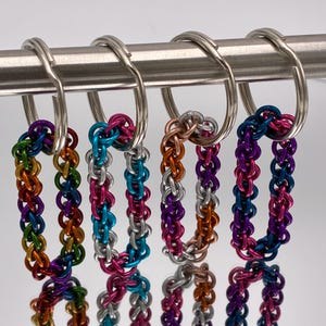 Może przedstawiać: Kolekcja kolorowych breloków z chainmaille. Każdy brelok ma inną kombinację kolorów, w tym tęczowy, różowy i srebrny, miedziany i fioletowy oraz niebieski i fioletowy. Breloki wiszą na srebrnych kółkach.