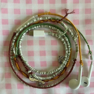 May include: Earbuds with a decorative cord wrapped in green, yellow, and maroon thread. The cord has silver charms, including a key and a leaf. The earbuds are white, and the charger is white with a USB-C connector.