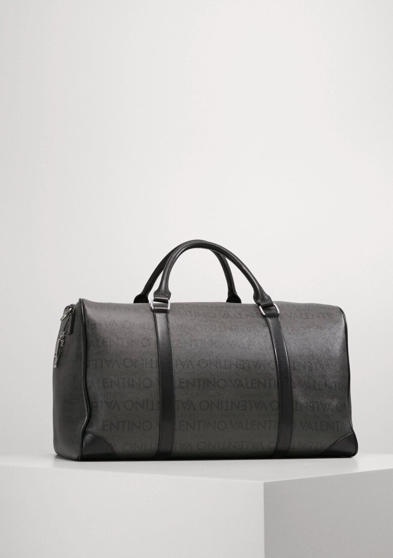 May include: A dark gray duffel bag with black handles and trim. The bag features the repeating text "VALENTINO" in a lighter gray pattern across its surface. The bag has a rectangular shape with a zipper closure and two top handles.