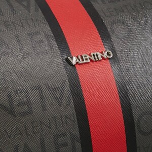 May include: Close-up of a textured gray fabric with a red stripe and a black stripe. The word "VALENTINO" is printed repeatedly on the gray fabric. A silver "VALENTINO" logo is centered on the red stripe.
