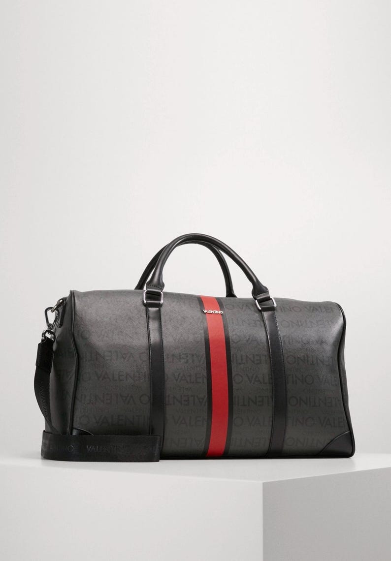 May include: A dark gray duffel bag with black handles and a shoulder strap. The bag features a red stripe down the center and the word "VALENTINO" printed repeatedly in a lighter gray.