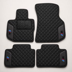 Può includere: Set di quattro tappetini neri per auto. Ogni tappetino presenta un motivo trapuntato a diamante e un logo con strisce blu, rosse e bianche. I tappetini sono progettati per i vani piedi di un veicolo.
