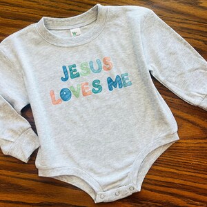 May include: A light gray baby bodysuit with long sleeves. The text "JESUS LOVES ME" is printed across the front in colorful letters. The bodysuit has a round neckline and snaps at the bottom.