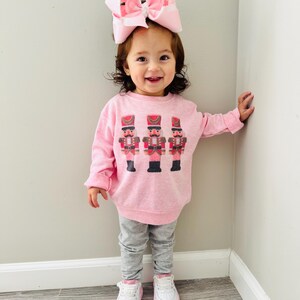 May include: A young child wearing a pink sweatshirt with a nutcracker graphic, gray leggings, and white and pink sneakers. A large pink bow adorns the child's hair. The child is smiling and standing near a wall.