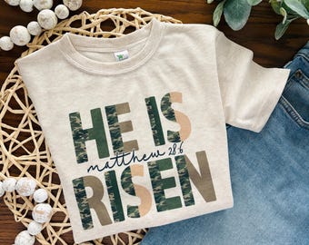 May include: Beige t-shirt with the text "HE IS RISEN" in green camouflage and tan letters. The text "Matthew 28:6" is written in a dark blue script. The shirt is folded on a woven mat with a pair of blue jeans.